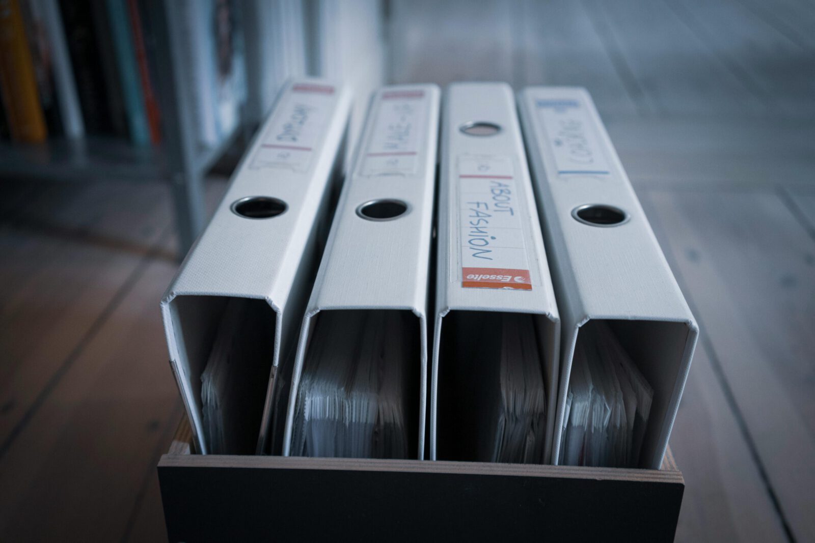 Four labeled office binders organized neatly on a wooden shelf in an indoor setting.